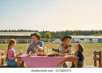 Young European Hipster Farm Family Having Lunch With Fresh Organic Products In Countryside Outdoors. Mother Feeding Son With Pepper. Father Showing Grapes To Daughter. Modern Farm Lifestyle. Sunny Day