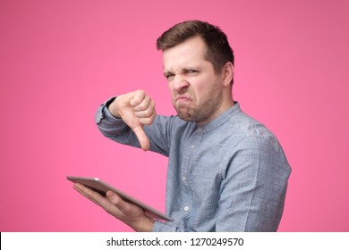 Young European Hipster Man Doing Bad Signal Showing Thumb Down Holding Tablet In Hands. He Got Bad News Or Read Something Negative In Internet.