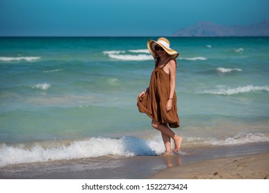 Young European lady in colorful bonnet hat and beautiful dress walking along picturesque wavy sea, she enjoying serenity around.