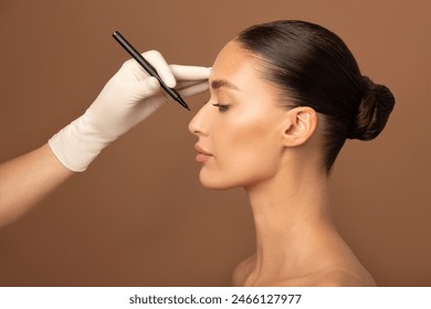 Young European lady getting prepared for plastic surgery procedures by doctor drawing contours on her nose, standing on beige background, side view - Powered by Shutterstock