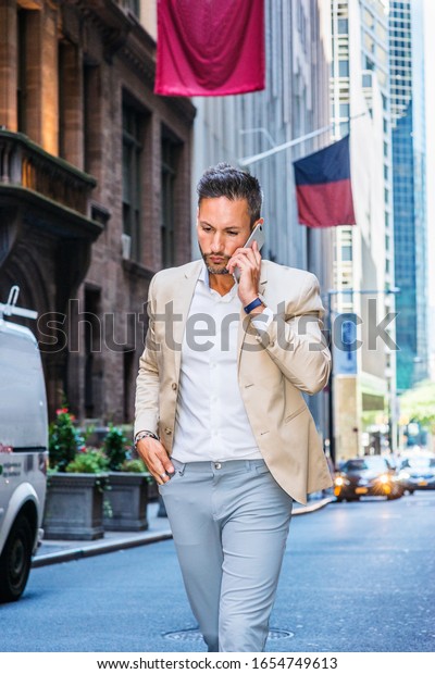 Young European Man with beard, little gray hair,\
traveling in New York, wearing beige blazer, white shirt, gray\
pants, walking on narrow street with high buildings in Manhattan,\
talking on cell phone.