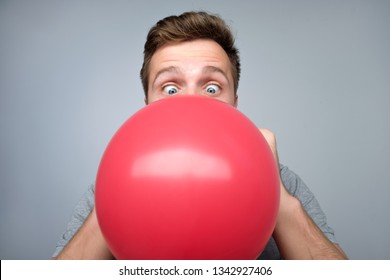 Young european man blowing up a red balloon preparing for party on gray background