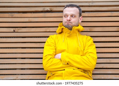 Young European Man With Sly Look, Beard Sitting Outside On Wooden Bench In Yellow Jacket With Crossed Hands, Blowing Cheeks, Looking Aside, Cunning Offended, Thinking Over Something, Keeping A Secret