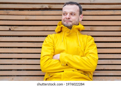 Young European Man With Sly Smile Sitting Outside On Wooden Bench In Bright Yellow Jacket With Crossed Hands, Thinking Over Idea, Plan, Looking Aside, Cunning Look, Dreaming Or Imagining Something