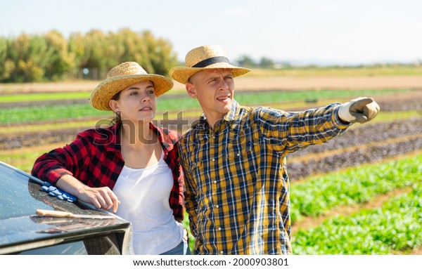 Young european man and woman\
farm workers talking near car on a farm field, pointing to\
something