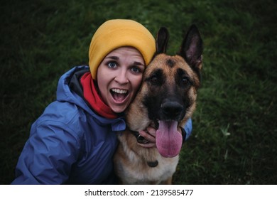 Young European white woman sits in green grass and hugs her German Shepherd dog top view. Have fun in park with furry human friend. Snow-white smile and tongue sticking out from joy.