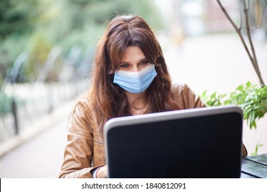 A Young European Woman In A Blue Protective Medical Mask Sits On The Street In A Cafe And Works Online Using A Laptop, Communication, Training And Video Communication Via The Internet, Social Distance