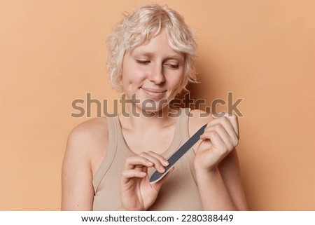 Young European woman with fair hair makes manicure fills her nails undergoes beauty treatments dressed in t shirt isolated over brown background takes care of herself smiles and has dimples on cheeks