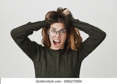 Young european woman in glasses and dark-green trendy sweater, screaming with angry and depressed expression, pulling her hair, isolated over white background. Girl can't stand her dormitory neighbor.
