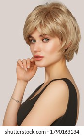 A Young European Woman In A Natural Looking Blonde Wig With Bangs Is Half-turned On The Gray Backdrop. The Lady Is Wearing A Black Tank Top And A Silver Bracelet With A Heart-shaped Charm. 