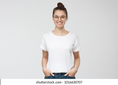 Young european woman standing with hands in pockets, wearing blank white tshirt with copy space for your logo or text, isolated on grey background