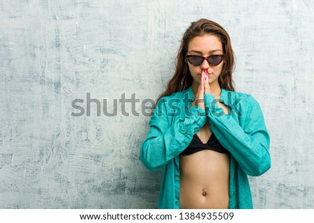 Young european woman wearing bikini holding hands in pray near mouth, feels confident.