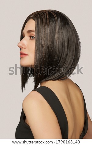 A young European woman is wearing a natural looking black wig with chestnut highlighting. The lady with blunt bob haircut is dressed in a black tank top and half-turned on the gray backdrop. 