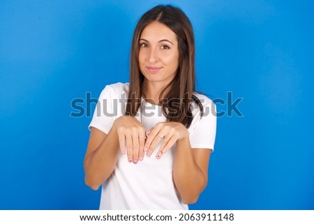 Young european woman wearing white T-shirt on blue background makes bunny paws and looks with innocent expression plays with her little kid