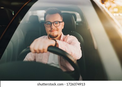 A young europen beautiful smiling man wearing glasses driving a car in the sity on sunset. View through the windshield 