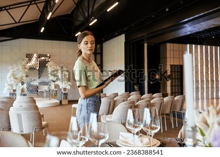 young event manager with clipboard looking at table with festive setting in modern event hall