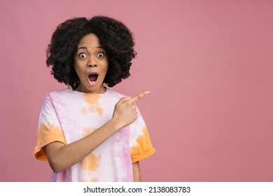 Young Excited African American Woman Looking At Camera Pointing Finger On Copy Space. Overjoyed Curly Haired Hipster Female Wearing Tie Dye T Shirt Isolated On Pink Background. Shopping, Sale Concept