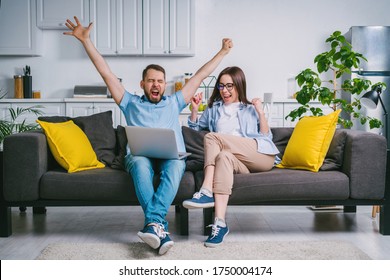 Young excited couple sitting at home on comfortable sofa watching soccer play live broadcast online on their laptop cheering for their favourite team happy with the score results.