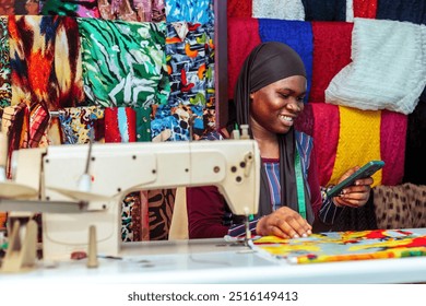 a young excited female African tailor inside a fashion clothing workshop using mobile phone while isolated over a colorful clothing background - Powered by Shutterstock