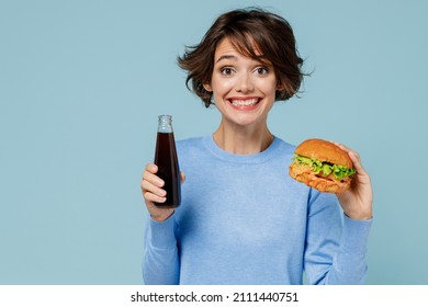 Young Excited Fun Cool Woman 20s Wearing Casual Sweater Look Camera Biting Eating Burger Drink Soda Cola Water Isolated On Plain Pastel Light Blue Background Studio. People Lifestyle Junk Food Concept