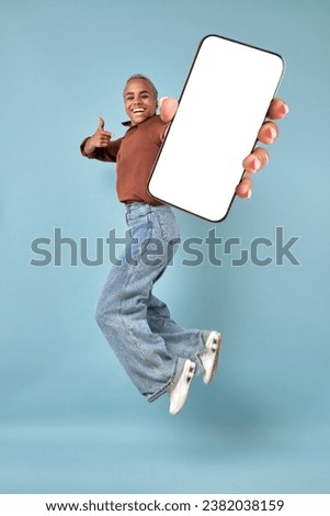 Young excited happy African American woman jumping up and showing off mobile phone and pointing finger at white screen of gadget with copy space for advertising application located in turquoise studio