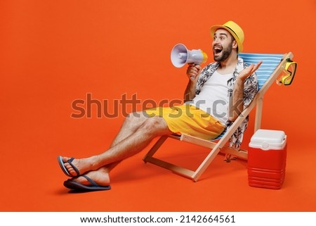 Young excited happy cool tourist man in beach shirt hat lie on deckchair near fridge scream in megaphone isolated on plain orange background studio portrait. Summer vacation sea rest sun tan concept.