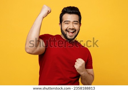 Young excited happy Indian man he wears red t-shirt casual clothes doing winner gesture celebrate clenching fists say yes isolated on plain yellow orange background studio portrait. Lifestyle concept