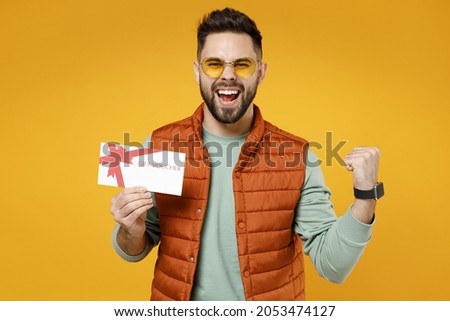 Young excited happy overjoyed caucasian man in orange vest mint sweatshirt glasses holding gift voucher flyer mock up do winner gesture clench fist celebrating isolated on yellow background studio.