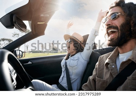 Young excited heterosexual caucasian couple driving a car. They are in vacation trip happy under sunlight. Girl rising arms in a cabrio and laughing a lot. People having fun and feeling freedom.