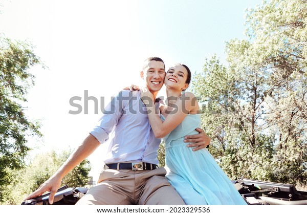 Young excited joyful couple, man\
and woman dressed festively while sitting on car roof, screaming\
and smiling from happiness while raising hands up in\
air