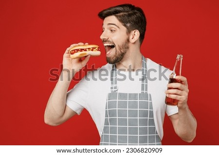Young excited male housewife housekeeper chef cook baker man wear grey apron hold in hand bottle drink beer eat hotdog biting open mouth isolated on plain red background studio. Cooking food concept