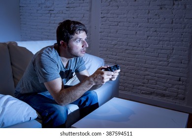 young excited man at home sitting on living room sofa playing video games using remote control joystick with freak intense face expression having fun in gaming addiction