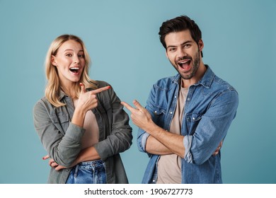 Young Excited Man And Woman Pointing Fingers At Each Other Isolated Over Blue Background