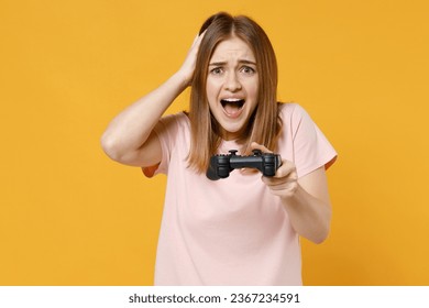 Young excited overjoyed disappointed displeased loser woman 20s wearing casual basic pastel pink t-shirt playing pc game with joystick console hold head isolated on yellow background studio portrait. - Shutterstock ID 2367234591