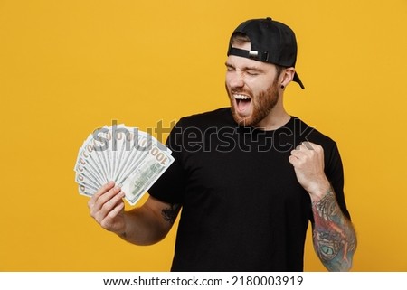 Young excited rich bearded tattooed man 20s he wears casual black t-shirt cap holding fan of cash money in dollar banknotes do winner gesture isolated on plain yellow wall background studio portrait.