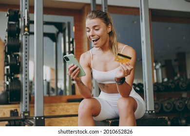 Young Excited Sportswoman Woman In White Sportswear Warm Up Train Sit Near Trainers Using Mobile Cell Phone Credit Bank Card For Online Shop Pay Gym Membership In Gym Indoors Workout Sport Concept.