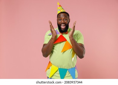 Young Excited Surprised Amazed Cool Fun Black Gay Man 20s In Green T-shirt Hat Cone Colorful Flags Celebrating Birthday Party Spread Hands Isolated On Plain Pastel Pink Background Studio Portrait.