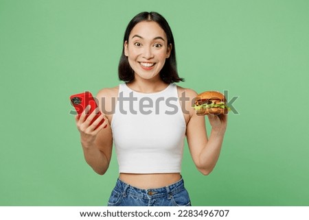Young excited woman wear white clothes hold eat burger use mobile cell phone application isolated on plain pastel light green background. Proper nutrition healthy fast food unhealthy choice concept