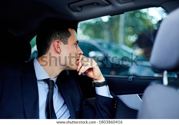 Young executive man traveling in\
the back seat of a car with a driver looking out the\
window