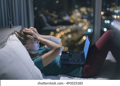 Young exhausted,depressed,concentrated woman lying in bed in bedroom with french windows in the dark at the lamp.Studies late at night.Staying up late. Overworking. Suffering from headache.