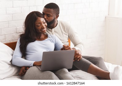 Young expecting woman and her loving husband using laptop while relaxing on bed at home, checking on furniture for baby room, copy space