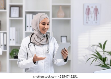 Young experienced female doctor in hijab using tablet computer for video call, female clinic worker consults patients remotely online, smiling contentedly inside clinic office. - Powered by Shutterstock