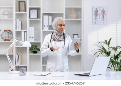 Young experienced female doctor in hijab using tablet computer for video call, female clinic worker consults patients remotely online, smiling contentedly inside clinic office. - Powered by Shutterstock