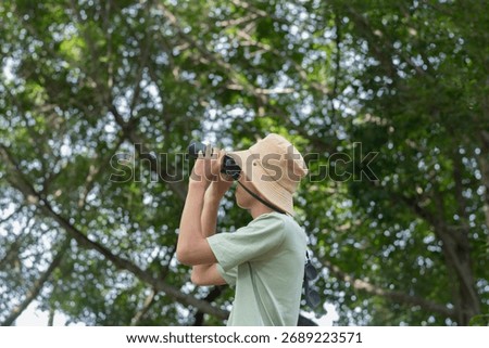 A young explorer is deeply engrossed in observing the tree canopy, looking upwards through his binoculars in the dense forest.