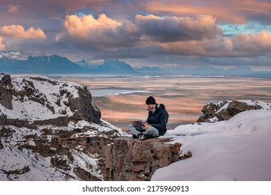 Young explorer using laptop while relaxing on snow covered cliff. Scenic view of volcanic landscape against cloudy sky. Male tourist using wireless technology during sunset.