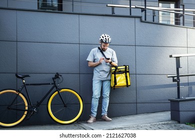 Young express food delivery courier with insulated bag on his side is looking at his phone near the parked bicycle.