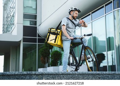 Young express food delivery courier riding bicycle with insulated bag behind his back is looking at his watch.