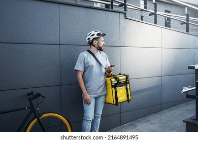 Young express food delivery courier with insulated bag on his side is holding phone near the parked bicycle.