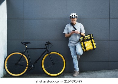 Young express food delivery courier with insulated bag on his side is looking at his phone near the parked bicycle.