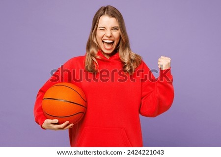 Young exultant jubilant fun woman fan wear red hoody cheer up support basketball sport team hold in hand ball do winner gesture isolated on plain purple color background studio. Sport leisure concept